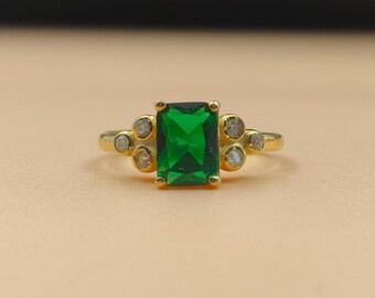 May include: A gold ring featuring a large, emerald-cut green gemstone as the centerpiece. The ring is accented with smaller, round, clear gemstones on each side of the main stone. The band is a simple, polished gold.