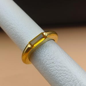 May include: A gold ring with a rectangular, yellow gemstone. The ring is set in a simple, polished gold band. The gemstone is a light yellow color and is set horizontally across the finger.