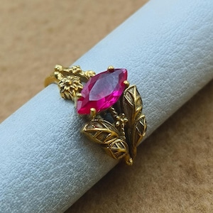 Può includere: Un anello dorato con una grande gemma magenta a taglio marquise. Il design dell'anello incorpora motivi floreali e a foglie, aggiungendo un elemento ornato e naturalistico al gioiello. L'anello è esposto su una superficie azzurra.