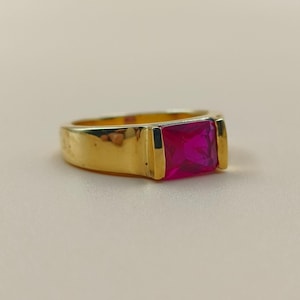 Puede incluir: Un anillo de oro con una piedra preciosa rectangular de color rosa brillante. El anillo tiene un diseño simple y clásico con un acabado pulido. La piedra preciosa está engastada en un bisel y el anillo parece ser de oro.