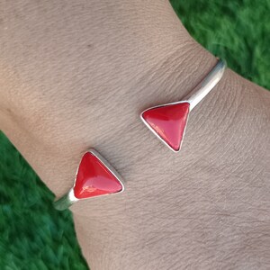 May include: A silver-toned open cuff bracelet featuring two polished, triangular red inlays at each end. The metal band is smooth and curved, designed to sit around the wrist with the two red triangular accents facing each other.