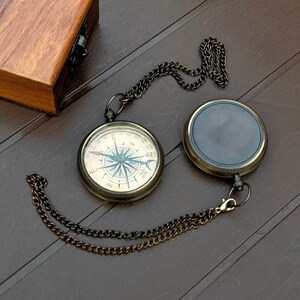 Peut inclure: Une boussole de poche ronde en laiton avec un cadran blanc, des points cardinaux noirs et une aiguille, fixée à une chaîne en métal sombre. À côté se trouvent un boîtier de boussole fermé assorti et une boîte en bois rectangulaire avec couvercle.
