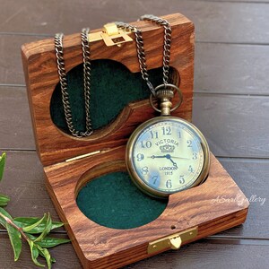 May include: A gold pocket watch with a black chain in a wooden box with a green felt lining. The watch face has black Roman numerals and the words 'VICTORIA' and 'LONDON 1915'.