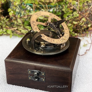 May include: A vintage brass sundial with a compass, set on a dark wooden box. The sundial features a circular design with Roman numerals and the text "Gilbert & Son London". The box has a metal clasp.