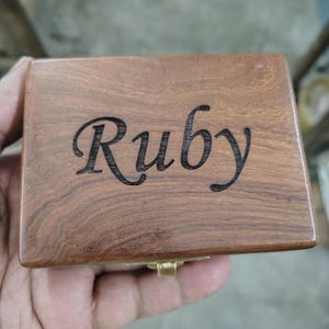 May include: A rectangular wooden box with a smooth, polished brown finish and visible wood grain. The name Ruby is engraved in a dark, cursive script across the center of the lid. A small, metallic gold-colored latch is visible at the front edge.