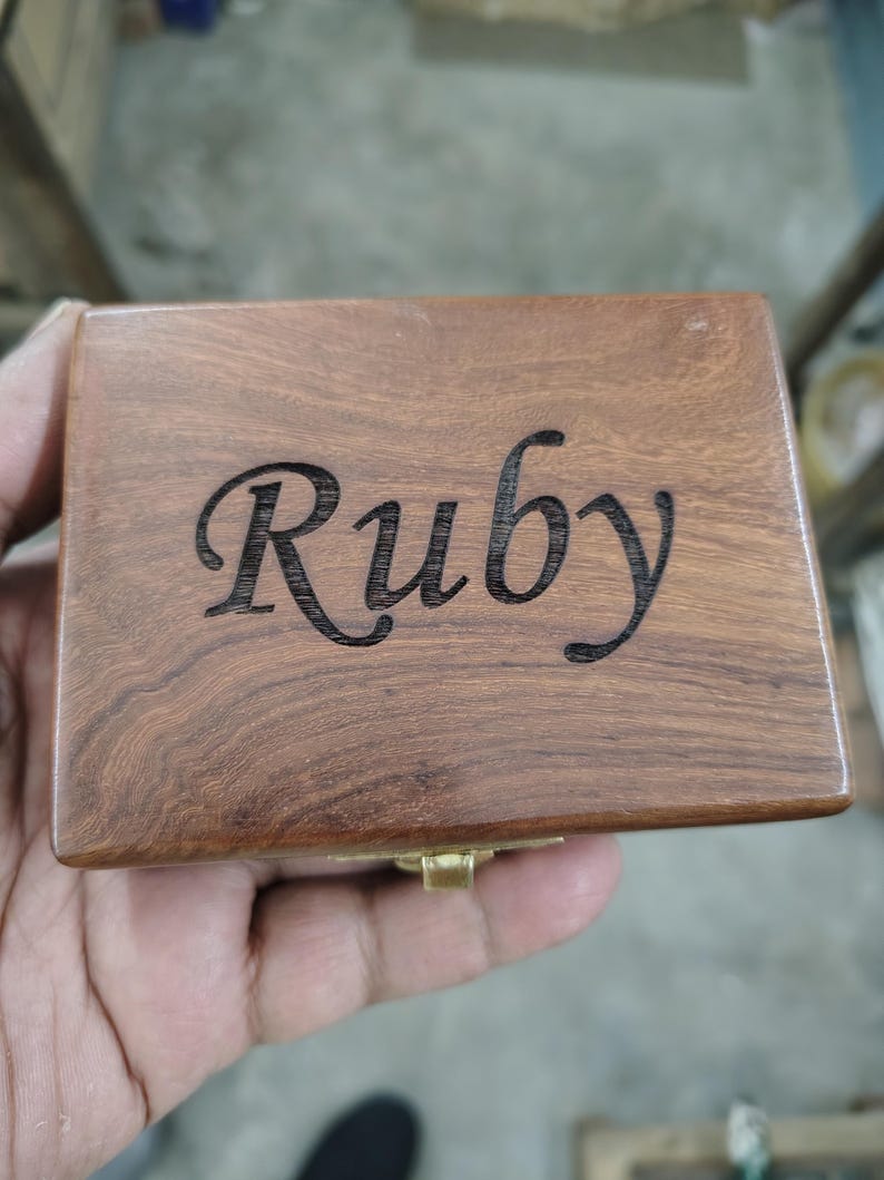 May include: A rectangular wooden box with a smooth, polished brown finish and visible wood grain. The name Ruby is engraved in a dark, cursive script across the center of the lid. A small, metallic gold-colored latch is visible at the front edge.
