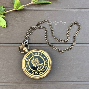 May include: A gold-toned pocket watch with a black face and a chain. The face of the watch has the words "Royal Navy London-1915" and a depiction of a crowned woman.