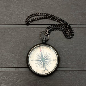 May include: A round, dark metal pocket compass featuring a cream-colored face with black cardinal directions, degree markings, and a needle with a red tip. The compass is attached to a matching dark metal chain with a circular link at the top.