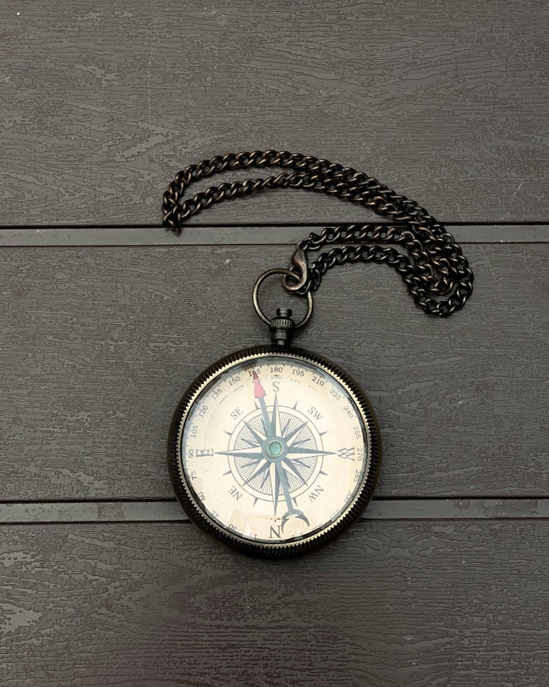 May include: A round, dark metal pocket compass featuring a cream-colored face with black cardinal directions, degree markings, and a needle with a red tip. The compass is attached to a matching dark metal chain with a circular link at the top.