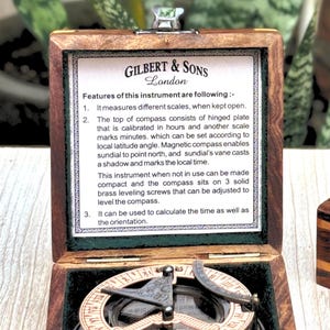 Może przedstawiać: Drewniane pudełko zawierające kompas magnetyczny i zegar słoneczny. Wewnętrzna strona wieczka posiada etykietę Gilbert & Sons London, opisującą funkcje takie jak zawiasowa płytka, regulacja szerokości geograficznej oraz trzy mosiężne śruby.