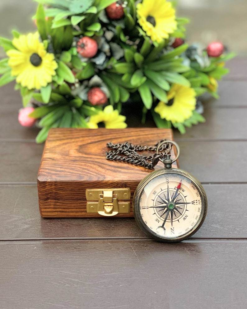 May include: A vintage-style compass with a chain, a green compass rose, and a red needle, resting on a wooden box with a brass latch. The compass is surrounded by a bouquet of yellow flowers.