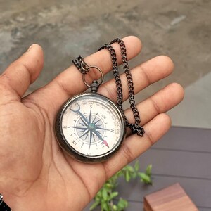 May include: A round, antique-style pocket compass with a dark metallic finish and a matching link chain. The compass face features a white background with black cardinal directions, a central star-shaped needle, and a red-tipped directional pointer.