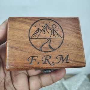 May include: A rectangular wooden box with a dark brown wood grain finish. The lid features a laser-engraved circular design depicting a mountain range, a winding path, and stars. Below the circular graphic, the letters F.R.M are engraved in a serif font.
