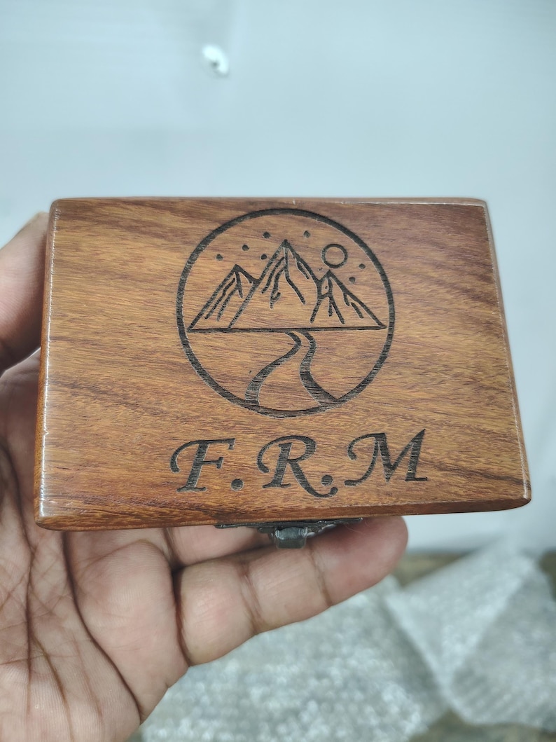 May include: A rectangular wooden box with a dark brown wood grain finish. The lid features a laser-engraved circular design depicting a mountain range, a winding path, and stars. Below the circular graphic, the letters F.R.M are engraved in a serif font.