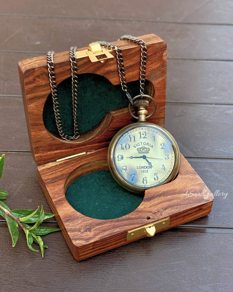 May include: A gold pocket watch with a black chain in a wooden box with a green felt lining. The watch face has black Roman numerals and the words 'VICTORIA' and 'LONDON 1915'.