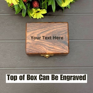 May include: A rectangular wooden box with a dark brown wood grain finish and a gold-colored metal latch. The lid features the engraved text "Your Text Here" centered on the surface. The box is positioned below a decorative arrangement of yellow flowers.