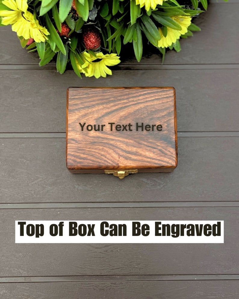 May include: A rectangular wooden box with a dark brown wood grain finish and a gold-colored metal latch. The lid features the engraved text "Your Text Here" centered on the surface. The box is positioned below a decorative arrangement of yellow flowers.