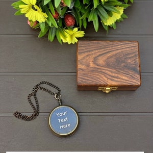 May include: A round antique-style compass with a dark metal chain and a blue face featuring the text Your Text Here, accompanied by a rectangular dark brown wooden box with a gold-toned metal latch, positioned near a bouquet of yellow flowers.
