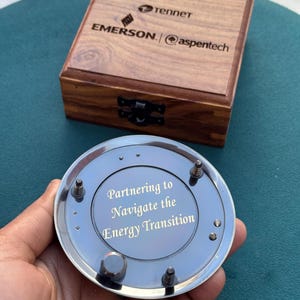 Może przedstawiać: Okrągły, srebrzysty metalowy przedmiot z wygrawerowanym napisem "Partnering to Navigate the Energy Transition", umieszczony przed kwadratowym drewnianym pudełkiem z logo Tennet, Emerson i aspentech na wieczku.