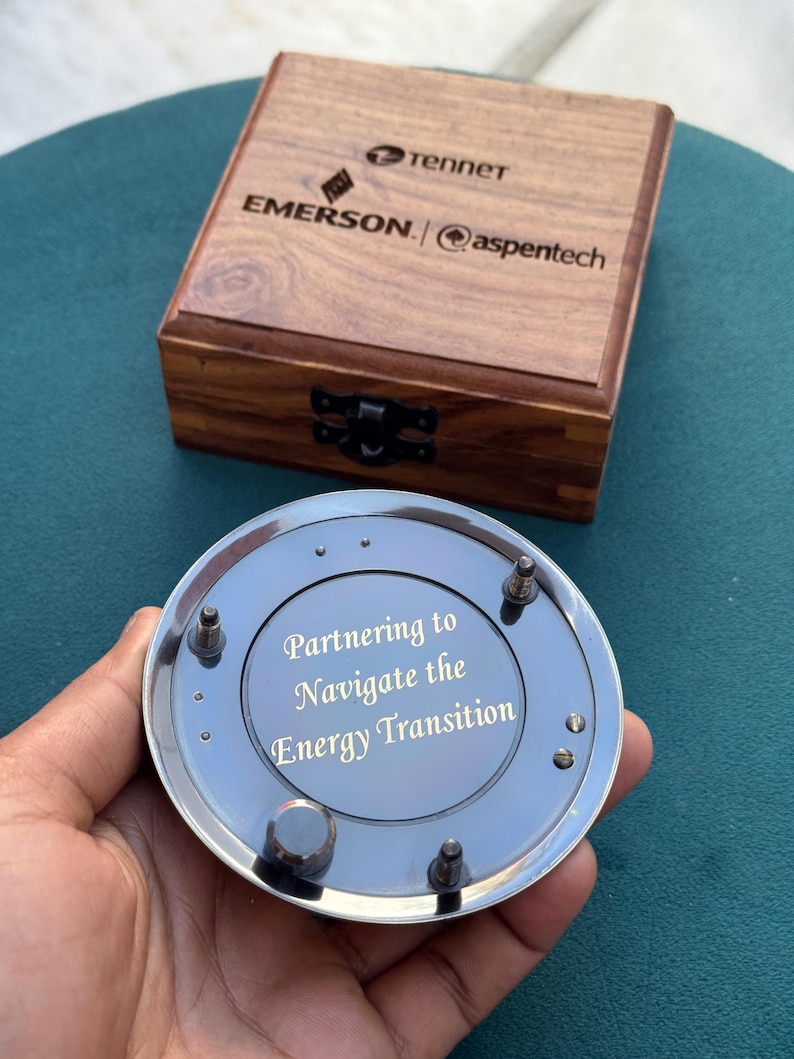 Może przedstawiać: Okrągły, srebrzysty metalowy przedmiot z wygrawerowanym napisem "Partnering to Navigate the Energy Transition", umieszczony przed kwadratowym drewnianym pudełkiem z logo Tennet, Emerson i aspentech na wieczku.