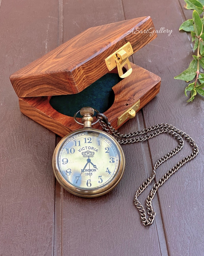 May include: A gold pocket watch with a black face and Roman numeral markings. The watch has a chain and is in a wooden box with a brass latch. The watch face says "Victoria" and "London 1915".