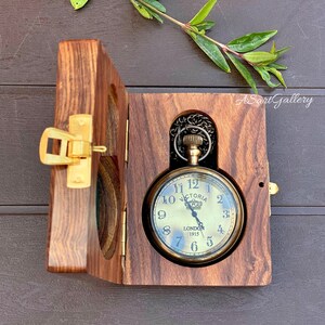 May include: A gold pocket watch with a black face and Roman numerals. The watch is in a wooden box with a hinged lid. The watch face says "Victoria London 1915".