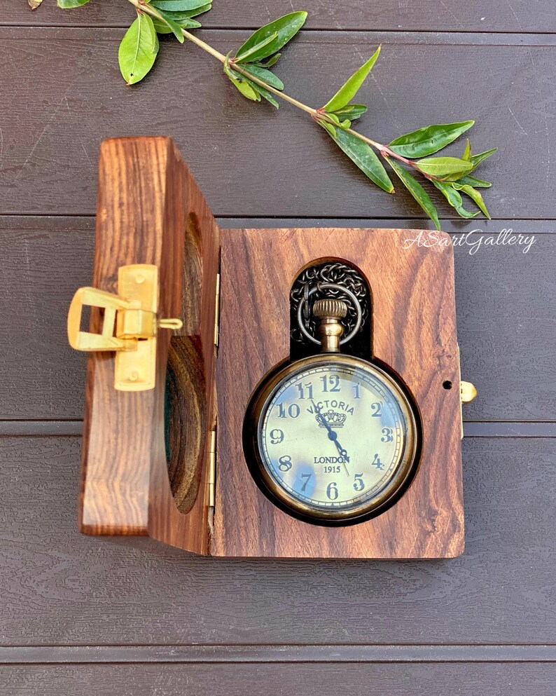 May include: A gold pocket watch with a black face and Roman numerals. The watch is in a wooden box with a hinged lid. The watch face says "Victoria London 1915".