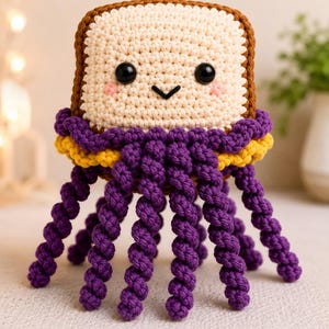 Peut inclure: Un jouet en peluche au crochet en forme de tranche de pain avec une croûte brune et un centre crème. Il présente des yeux de sécurité noirs, une petite bouche noire brodée et des joues roses. Sous le pain se trouvent des tentacules en laine violette et jaune.
