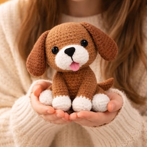 Könnte beinhalten: Handgehäkelter Welpe mit braunen und weißen Details. Der Hund hat schwarze Augen, eine rosa Zunge und Schlappohren. Der Hund wird in den Händen gehalten.