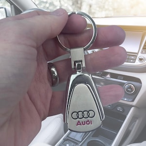 May include: Silver Audi keyring featuring the Audi logo and brand name in pink. The keyring is held in a hand, with a car interior visible in the background.