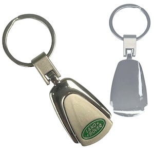 May include: A silver Land Rover keyring with a key ring. The keyring features a polished, teardrop-shaped design with the Land Rover logo in green and white. The keyring is made of metal.