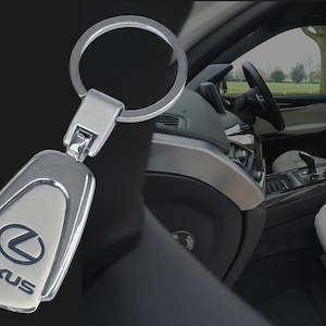 May include: A silver Lexus keyring with a polished finish. The keyring features the Lexus logo and text on a silver background. The keyring is attached to a silver ring. The background shows the interior of a car.