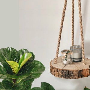 May include: A wooden hanging shelf with a natural bark finish. The shelf is suspended by three ropes and holds two glass candle holders. One candle holder is clear and the other is frosted.