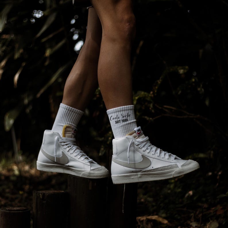 May include: White high-top sneakers with a gray swoosh logo and a cream-colored sole. The sneakers are paired with white socks that have the text "Coole Socke Seit 1989". The shoes are on a wooden surface.