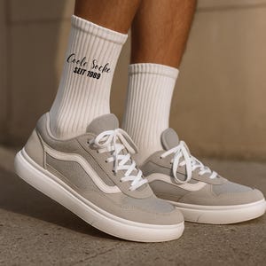 May include: A pair of light gray sneakers with white laces and a white sole. The shoes are paired with white ribbed socks. The socks have the text "Coole Socke SEIT 1989" printed on them.