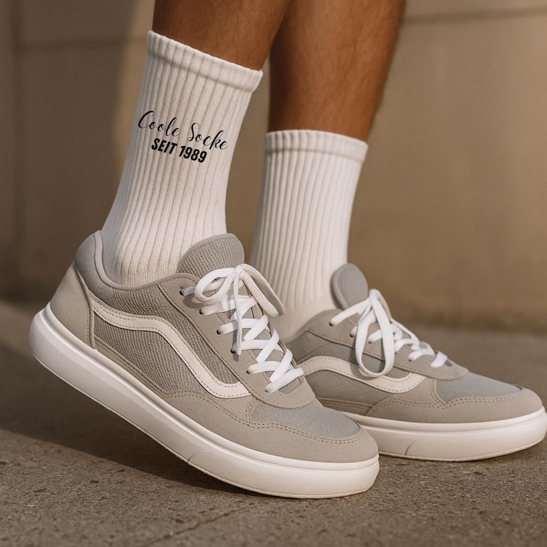 May include: A pair of light gray sneakers with white laces and a white sole. The shoes are paired with white ribbed socks. The socks have the text "Coole Socke SEIT 1989" printed on them.