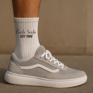May include: A pair of gray sneakers with white laces and a white side stripe. The sneakers are paired with white ribbed socks that have the text "Coole Socke SEIT 1989" printed on them. The shoes have a white sole.