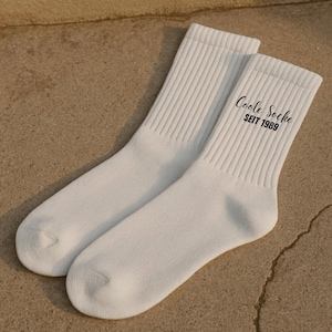 May include: A pair of white crew socks with ribbed cuffs. The text "Coole Socke SEIT 1989" is printed in black on the side of one sock. The socks are made of a soft, comfortable material.