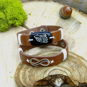 May include: Brown leather bracelet with a white leather accent and a silver infinity symbol charm. The bracelet also features a black oval charm with a white wolf design.