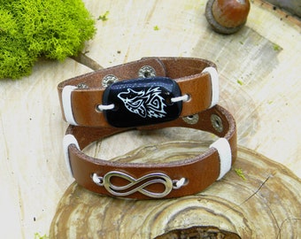 May include: Brown leather bracelet with a white leather accent and a silver infinity symbol charm. The bracelet also features a black oval charm with a white wolf design.