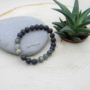 May include: A black lava rock and green agate beaded bracelet with silver spacers. The bracelet is lying on a light grey stone and a wooden surface.