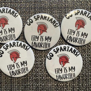 May include: Five white round buttons with the text "GO SPARTANS!" and "LILY IS MY FAVORITE!!" in black. Each button features a red Spartan helmet graphic. The buttons are arranged on a textured surface.