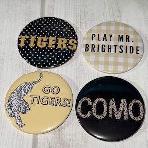 May include: Four round buttons with text and designs. One button has "TIGERS" in gold letters on a black background with white polka dots. Another says "PLAY MR. BRIGHTSIDE" on a yellow and white checkered background. A third says "GO TIGERS!" with a tiger illustration. The last says "COMO".