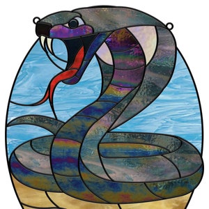 Op de afbeelding: Een glas-in-loodraam met een afbeelding van een cobra met iriserende blauwe, paarse en groene schubben. De cobra is opgerold met zijn kop omhoog en zijn bek open, waardoor een rode tong zichtbaar is. De achtergrond is een blauwe lucht met een gouden zandduin.