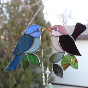 May include: A stained glass suncatcher featuring two birds perched on a branch with green leaves. The birds are blue and pink with black accents. The suncatcher has a silver chain and beads hanging from the bottom.