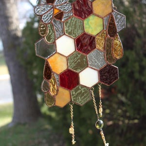 May include: A stained glass honeycomb with a bee hanging from the top. The honeycomb is made of various colors, including red, green, yellow, and white. The bee is made of silver and black glass. The honeycomb is hanging from a chain with gold beads.