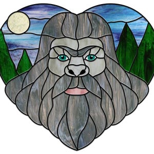 Peut inclure: Une œuvre d'art en vitrail en forme de cœur représentant une créature ressemblant à Bigfoot avec une barbe grise et des yeux bleus. L'arrière-plan présente un ciel bleu avec une lune blanche et des arbres verts.