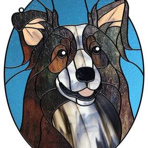 May include: Stained glass artwork depicting a dog's face, featuring brown, black, white, and blue glass pieces. The dog's face is oval-shaped with a blue background. The artwork includes a metal loop for hanging.
