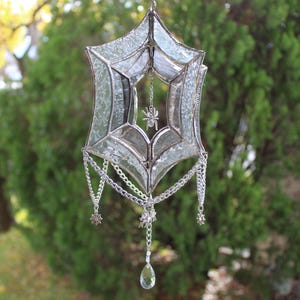 May include: A silver stained glass spider web suncatcher with a dangling crystal and silver chains. The web is made of clear glass with a spider in the center.