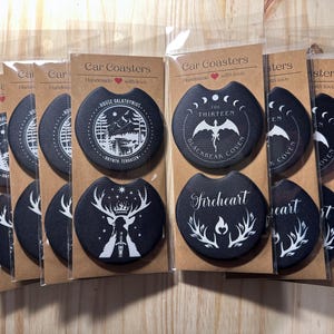 May include: A set of 10 black car coasters featuring white designs. The designs include a stag, a dragon, a moon, and a tree. The coasters are handmade with love and are ideal for adding a touch of whimsy to your car.